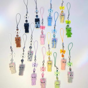 May include: A collection of colorful phone charms featuring small, fuzzy cat and frog figurines. Each charm has a loop for attachment, adorned with beads and decorative bows in various colors, including pink, blue, and yellow.