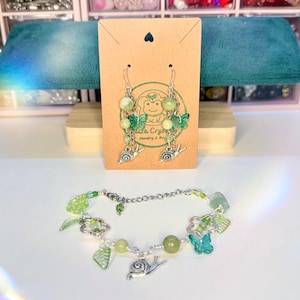 May include: A set of handmade green beaded earrings and bracelet. The earrings and bracelet feature green beads, butterfly charms, leaf charms, and silver snail charms. The earrings are displayed on a brown card with the text "Liza Crystal Jewelry & Kels".