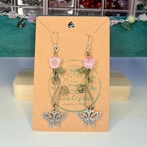 Könnte beinhalten: Ohrringe zum Aufhängen mit rosa tulpenförmigen Perlen, grünen Blattdetails und silbernen Schmetterlingsanhängern. Die Ohrringe hängen an silbernen Haken und haben filigrane Drahtarbeiten. Im Hintergrund ist eine Karte mit dem Text "Eliza Crystal Jewelry & More" zu sehen.
