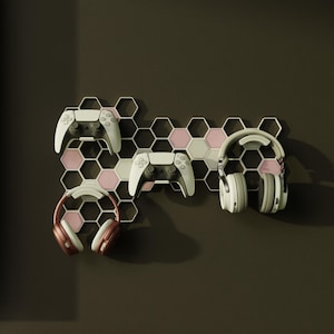 May include: A white and pink honeycomb-shaped wall mount holding two white game controllers and two pairs of headphones. The headphones are white and pink, and the controllers are white. The wall mount is against a dark brown wall.