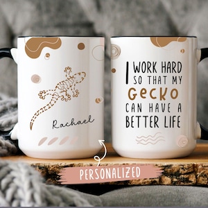 May include: White ceramic mug with a black handle. The mug has a brown gecko illustration and the text "Rachael" on one side. The other side has the text "I work hard so that my Gecko can have a better life".