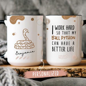 May include: Two white ceramic mugs with black handles. The mug on the left has a brown snake illustration and the text "Benjamin". The mug on the right has the text "I work hard so that my Ball Python can have a better life". Both mugs have a black rim and a personalized label.