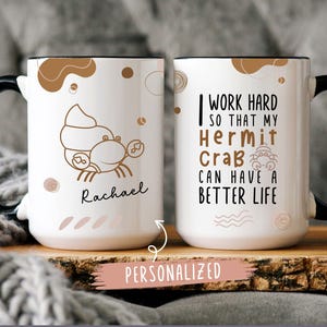 May include: White ceramic mug with a black handle. The mug has a brown hermit crab illustration and the text "I work hard so that my hermit crab can have a better life". The mug also has a brown abstract design and the name "Rachael" written on it.