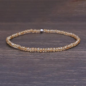 Könnte beinhalten: Ein zartes Armband aus kleinen, facettierten, bernsteinfarbenen Edelsteinen. Das Armband ist mit einem silberfarbenen Verschluss versehen. Die Edelsteine haben einen warmen, goldenen Farbton und liegen auf einer dunklen Holzoberfläche.