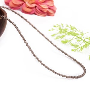 Op de afbeelding: Een delicate ketting gemaakt met kleine, donkergrijze edelsteen kralen. De ketting wordt getoond op een wit oppervlak met een roze bloem en groen gebladerte op de achtergrond.