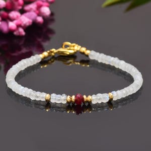 Könnte beinhalten: Ein zartes Armband aus weißen Mondsteinperlen und einer einzelnen roten Rubinperle. Das Armband ist mit goldfarbenen Zwischenstücken verziert.