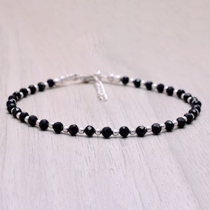 Può includere: Braccialetto alla caviglia con piccole perline nere sfaccettate e perline color argento. La delicata catena e la chiusura sono visibili. Il braccialetto è esposto su una superficie di legno chiaro.