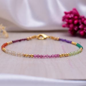 May include: A delicate bracelet with a gold clasp and a rainbow of gemstone beads. The beads are arranged in a graduated pattern, starting with orange, then light brown, pink, purple, and green.