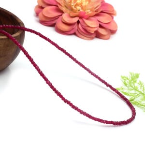 May include: A necklace made of small, square-shaped, ruby red beads. The necklace is lying on a white surface.