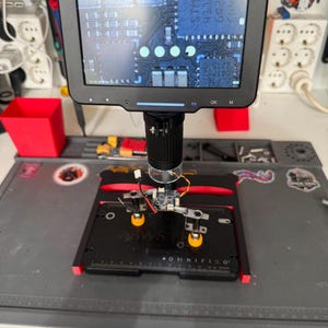 Peut inclure: Un microscope numérique avec un écran rectangulaire noir affichant un circuit imprimé agrandi. L'appareil comprend un support vertical, une lentille et une plaque de base noire marquée OMNIFIXO avec des pinces métalliques réglables.