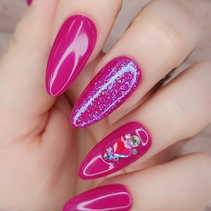 Puede incluir: Una mano que muestra uñas artificiales largas de color magenta. Una uña está cubierta de purpurina y otra presenta un diseño de joya en forma de corazón. Las uñas son de un color sólido y brillante, y el efecto general es glamuroso.