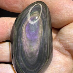May include: A polished, oval gemstone displaying a swirling pattern of dark grey, purple, and iridescent green hues. The stone has a glossy finish and a white, oval inclusion near the top. The gemstone is held in a hand.