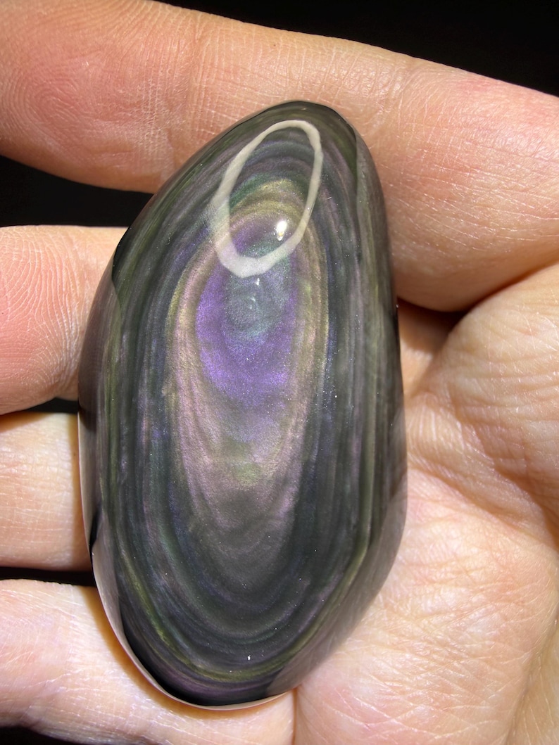 May include: A polished, oval gemstone displaying a swirling pattern of dark grey, purple, and iridescent green hues. The stone has a glossy finish and a white, oval inclusion near the top. The gemstone is held in a hand.