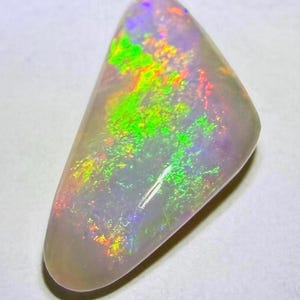 May include: A triangular-shaped polished opal gemstone with a translucent, milky white base. The surface displays vibrant flashes of neon green, bright orange, and subtle purple iridescence that shift across the smooth, rounded stone surface.