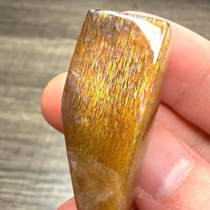 May include: A polished, elongated sunstone cabochon featuring a warm golden-brown hue with distinct, shimmering metallic inclusions that create a reflective, fibrous texture across the surface of the smooth, translucent gemstone.