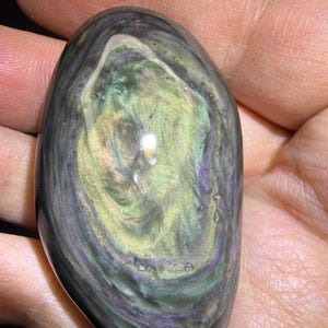 May include: An oval gemstone, polished to a high sheen, displaying a mix of colours including green, purple, and black. The stone is held in a hand, and the swirling patterns create an iridescent effect. The central area is a lighter green.