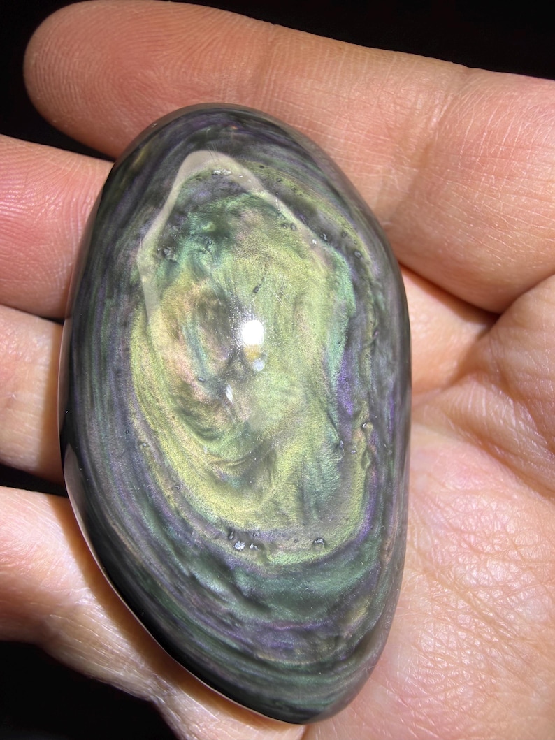 May include: An oval gemstone, polished to a high sheen, displaying a mix of colours including green, purple, and black. The stone is held in a hand, and the swirling patterns create an iridescent effect. The central area is a lighter green.