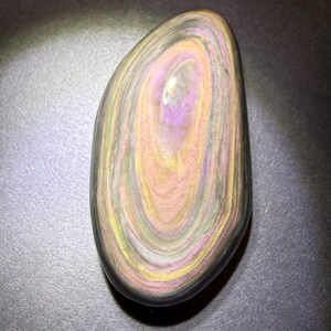 May include: A polished, oval gemstone with a swirling pattern of colours. The stone features bands of purple, yellow, and peach hues, with a dark outer edge. The gemstone is smooth and reflective, with a natural, organic appearance.