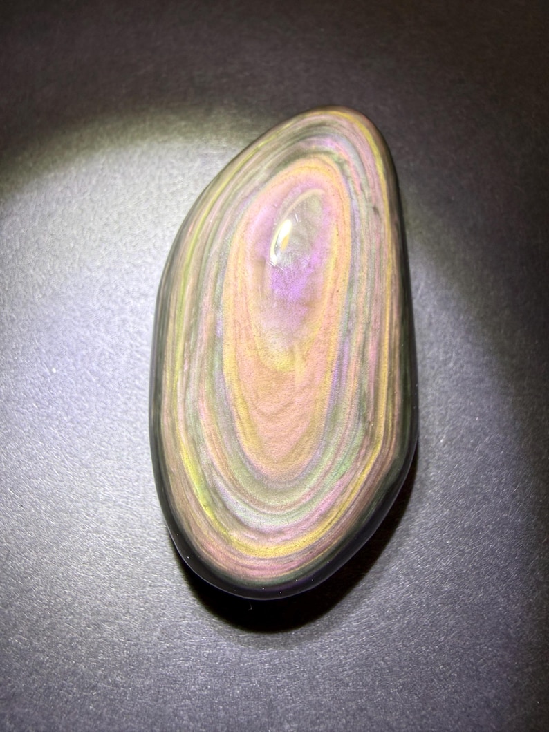 May include: A polished, oval gemstone with a swirling pattern of colours. The stone features bands of purple, yellow, and peach hues, with a dark outer edge. The gemstone is smooth and reflective, with a natural, organic appearance.