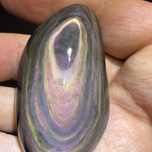 May include: A polished, oval obsidian stone with swirling bands of colour. The stone has a central eye-like pattern with shades of purple, pink, gold, and green. The surface is smooth and reflective, highlighting the natural beauty of the gemstone.