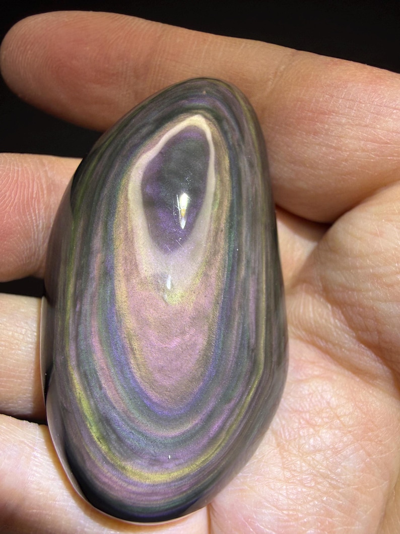 May include: A polished, oval obsidian stone with swirling bands of colour. The stone has a central eye-like pattern with shades of purple, pink, gold, and green. The surface is smooth and reflective, highlighting the natural beauty of the gemstone.