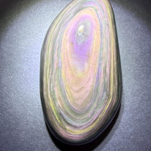 May include: A polished, oval-shaped rainbow obsidian stone. The stone features concentric bands of colour, including purple, yellow, green, and black. The surface is smooth and reflective, with a subtle sheen. The stone is a natural, decorative item.