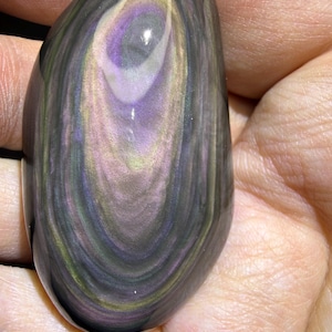 May include: A polished, oval-shaped obsidian stone with swirling patterns of purple, grey, yellow, and green. The stone is held in a hand, showcasing its smooth, reflective surface. The natural stone has a unique, eye-like design.