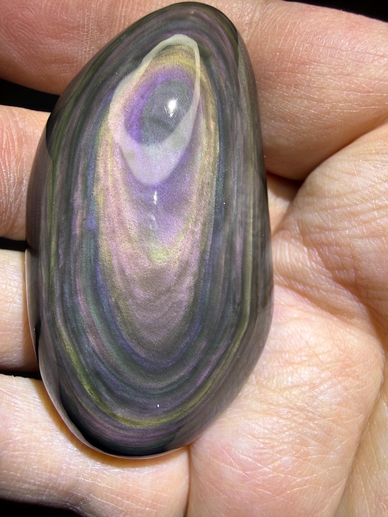 May include: A polished, oval-shaped obsidian stone with swirling patterns of purple, grey, yellow, and green. The stone is held in a hand, showcasing its smooth, reflective surface. The natural stone has a unique, eye-like design.