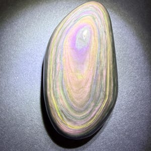 May include: A polished, oval-shaped stone with a swirling pattern of colours. The stone features bands of pink, yellow, green, and black, creating a unique, iridescent effect. The stone is smooth and appears to be a natural gemstone.