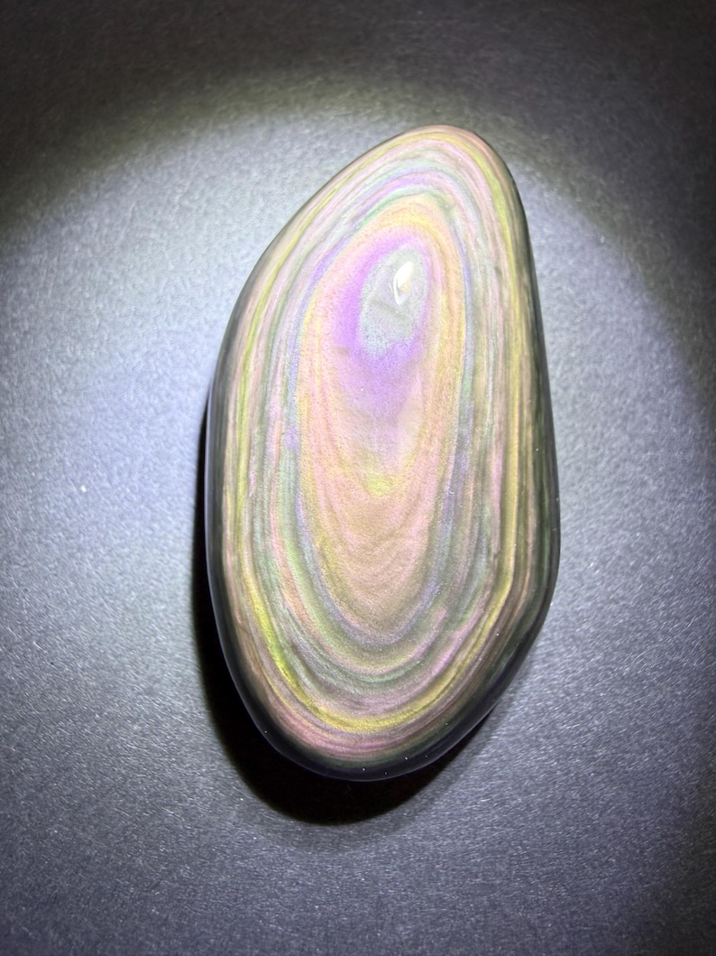 May include: A polished, oval-shaped stone with a swirling pattern of colours. The stone features bands of pink, yellow, green, and black, creating a unique, iridescent effect. The stone is smooth and appears to be a natural gemstone.