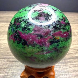 May include: A polished spherical stone featuring a vibrant mix of deep green, magenta, and black mineral inclusions. The sphere rests on a small, carved wooden tripod stand, showcasing a smooth, reflective surface with natural color variations.