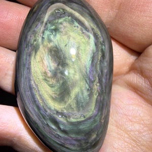 May include: An oval obsidian stone, polished to a high sheen, displaying swirling patterns of green, gold, and purple. The stone's surface reflects light, creating a shimmering effect. The dark base colour contrasts with the lighter, swirling patterns, highlighting the natural beauty of the gemstone.