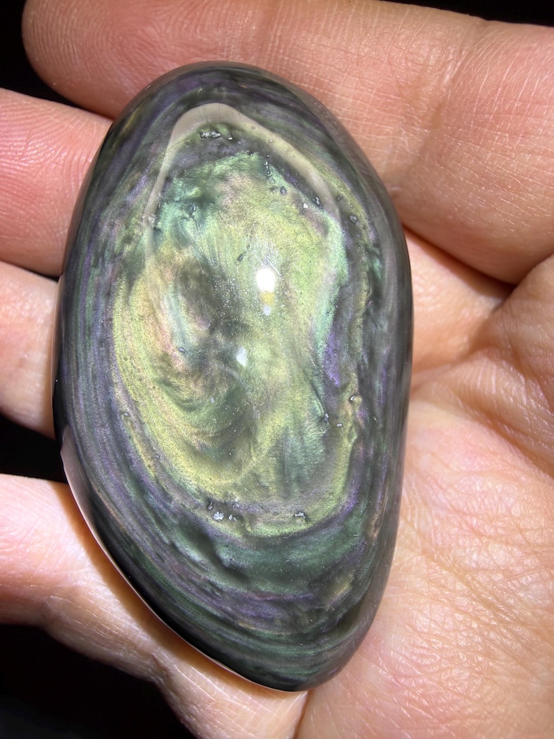 May include: An oval obsidian stone, polished to a high sheen, displaying swirling patterns of green, gold, and purple. The stone's surface reflects light, creating a shimmering effect. The dark base colour contrasts with the lighter, swirling patterns, highlighting the natural beauty of the gemstone.
