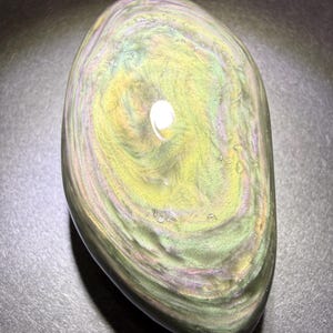 May include: An oval-shaped iridescent obsidian stone with polished surfaces. The stone features swirling patterns in green, yellow, and purple. The glossy finish reflects light, creating a shimmering effect. A natural, decorative item, ideal for display or collection.