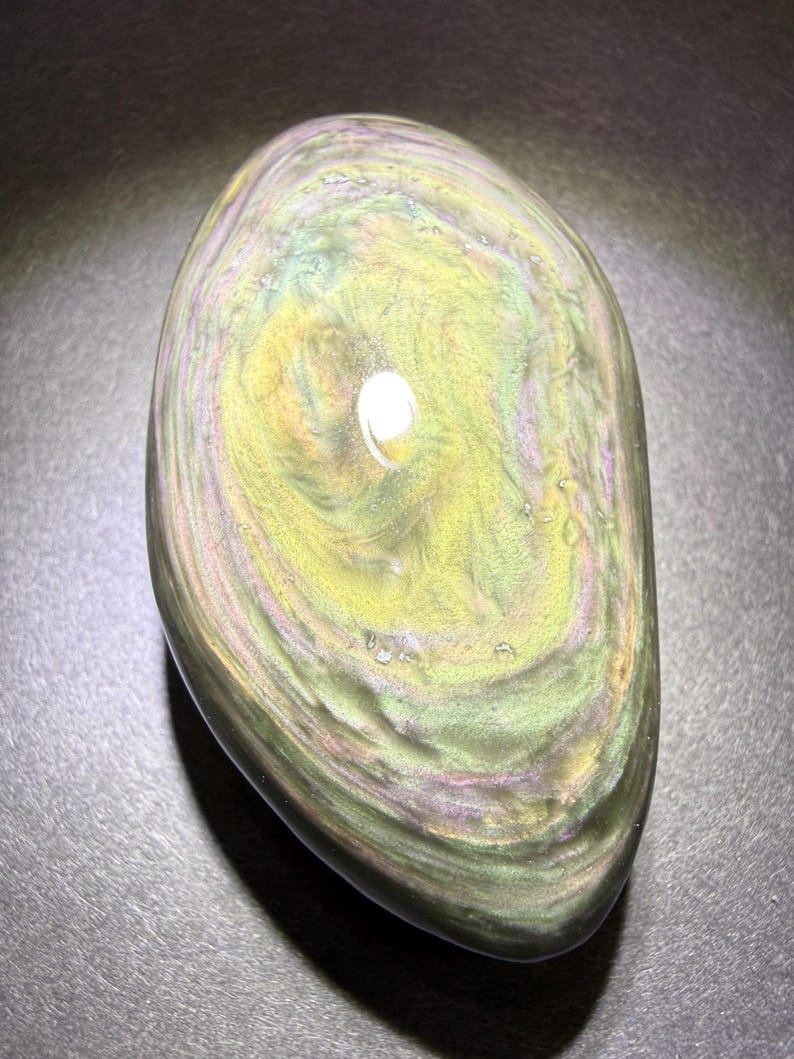 May include: An oval-shaped iridescent obsidian stone with polished surfaces. The stone features swirling patterns in green, yellow, and purple. The glossy finish reflects light, creating a shimmering effect. A natural, decorative item, ideal for display or collection.