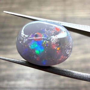 May include: A smooth, oval-shaped opal gemstone with a translucent, pale grey base color. The stone features vibrant, multi-colored flashes of red, green, and blue light throughout its surface, held securely by a pair of metal tweezers.