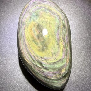 May include: A polished, oval-shaped rainbow obsidian stone. The stone displays swirling patterns of green, yellow, and purple hues. The surface is smooth and reflective, with a glossy finish.