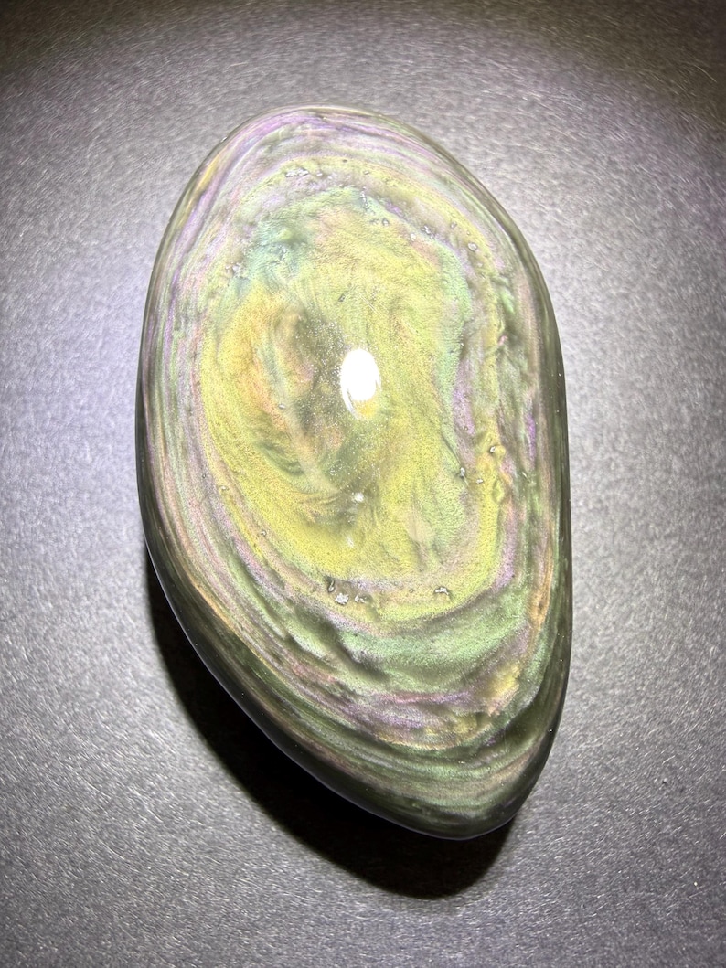 May include: A polished, oval-shaped rainbow obsidian stone. The stone displays swirling patterns of green, yellow, and purple hues. The surface is smooth and reflective, with a glossy finish.