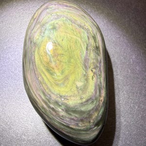 May include: A polished, oval gemstone displaying a swirling pattern of green, gold, and purple tones. The stone has a smooth, glossy surface and a dark, nearly black, base colour. The gemstone is a natural, unique piece.