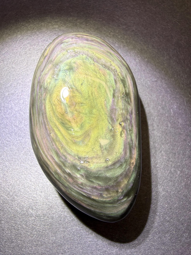 May include: A polished, oval gemstone displaying a swirling pattern of green, gold, and purple tones. The stone has a smooth, glossy surface and a dark, nearly black, base colour. The gemstone is a natural, unique piece.