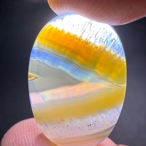 May include: An oval-shaped gemstone with layers of yellow, orange, and blue hues. The stone is held between two fingers, and the light shines through the translucent material, highlighting the color variations and patterns within the stone.