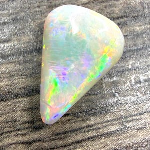 May include: A polished, teardrop-shaped white opal gemstone featuring a translucent base with vibrant flashes of green, yellow, orange, and purple play-of-color throughout its surface, showcasing a smooth, rounded, and asymmetrical cabochon cut.