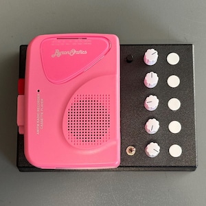 May include: A pink cassette player with the text "AM/FM RADIO RECORDER CASSETTE PLAYER" and the logo "ByronStatics" on the front. The player is sitting on a black box with four white knobs and four white circles.