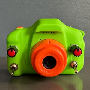 Op de afbeelding: Een felgroene speelgoedcamera met een oranje lens en rode accenten. De camera heeft twee zwarte draaiknoppen met rode centra en een zilveren schakelaar. Ontworpen voor kinderen.