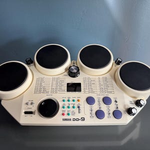 Puede incluir: Batería electrónica Yamaha DD-9 con cinco pads de batería negros y varias perillas de control. La unidad blanquecina presenta una pequeña pantalla y botones para estilo, tempo y efectos de sonido. El texto "Yamaha DD-9" está impreso en la parte delantera.