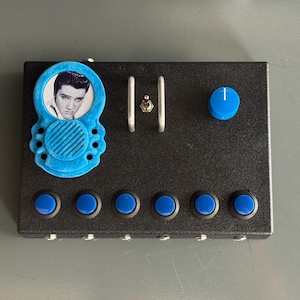 May include: Black rectangular electronic device with blue accents. Features a blue Elvis Presley portrait, six blue buttons, a blue knob, and a silver toggle switch. The device has a textured surface and appears to be custom-made.