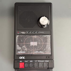 Może przedstawiać: Czarny radioodtwarzacz FM i odtwarzacz kaset QFX. Radio posiada głośnik, srebrne pokrętło i przezroczyste plastikowe drzwiczki na kasetę. Radio posiada przyciski do nagrywania, odtwarzania, przewijania do tyłu, szybkiego przewijania do przodu, zatrzymania/wysunięcia i pauzy. Tekst "FM RADIO" i "QFX."