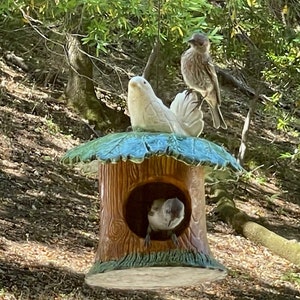 May include: A ceramic birdhouse shaped like a tree trunk with a green leafy roof. A small bird is perched on the roof, and another bird is peeking out of the opening.