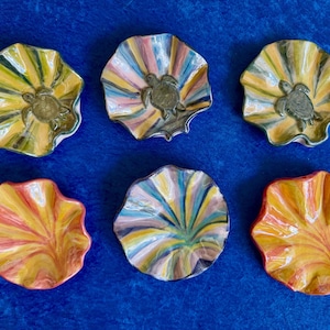 May include: Six decorative ceramic dishes with a seashell shape and a turtle design. The dishes are painted in various colors, including yellow, orange, pink, blue, and green. The dishes are arranged on a blue surface.