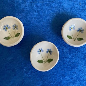 May include: Three small, round, white ceramic dishes. Each dish features a hand-painted design of blue flowers with green leaves. The dishes are arranged on a blue surface, showcasing a charming, handcrafted aesthetic.
